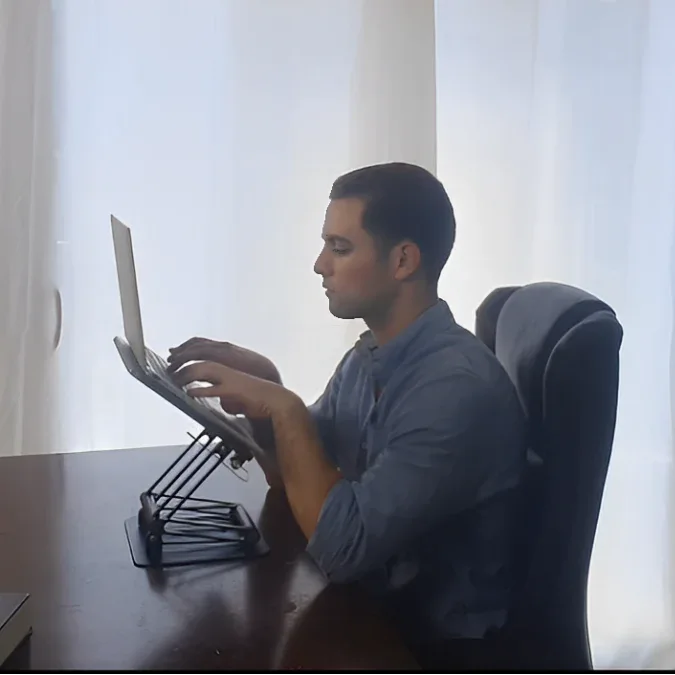 Man working on a laptop elevated on an adjustable stand with upright posture at a desk, promoting comfortable screen viewing during long work sessions